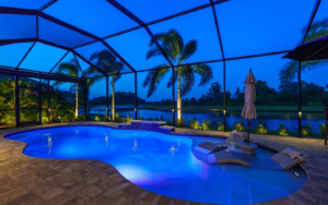 Screened backyard pool at dusk with blue lighting, palm trees, and a lake view beyond the enclosure.