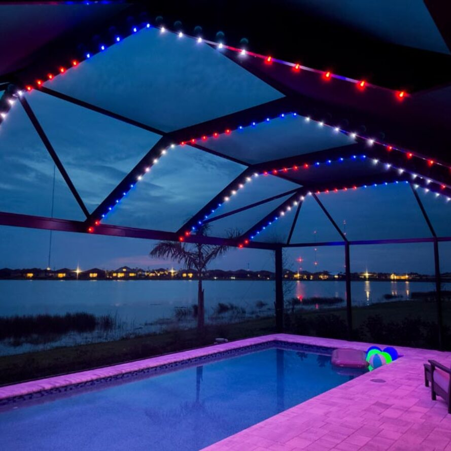 Color-changing LED puck lights outlining the large beams of a waterfront pool cage at dusk